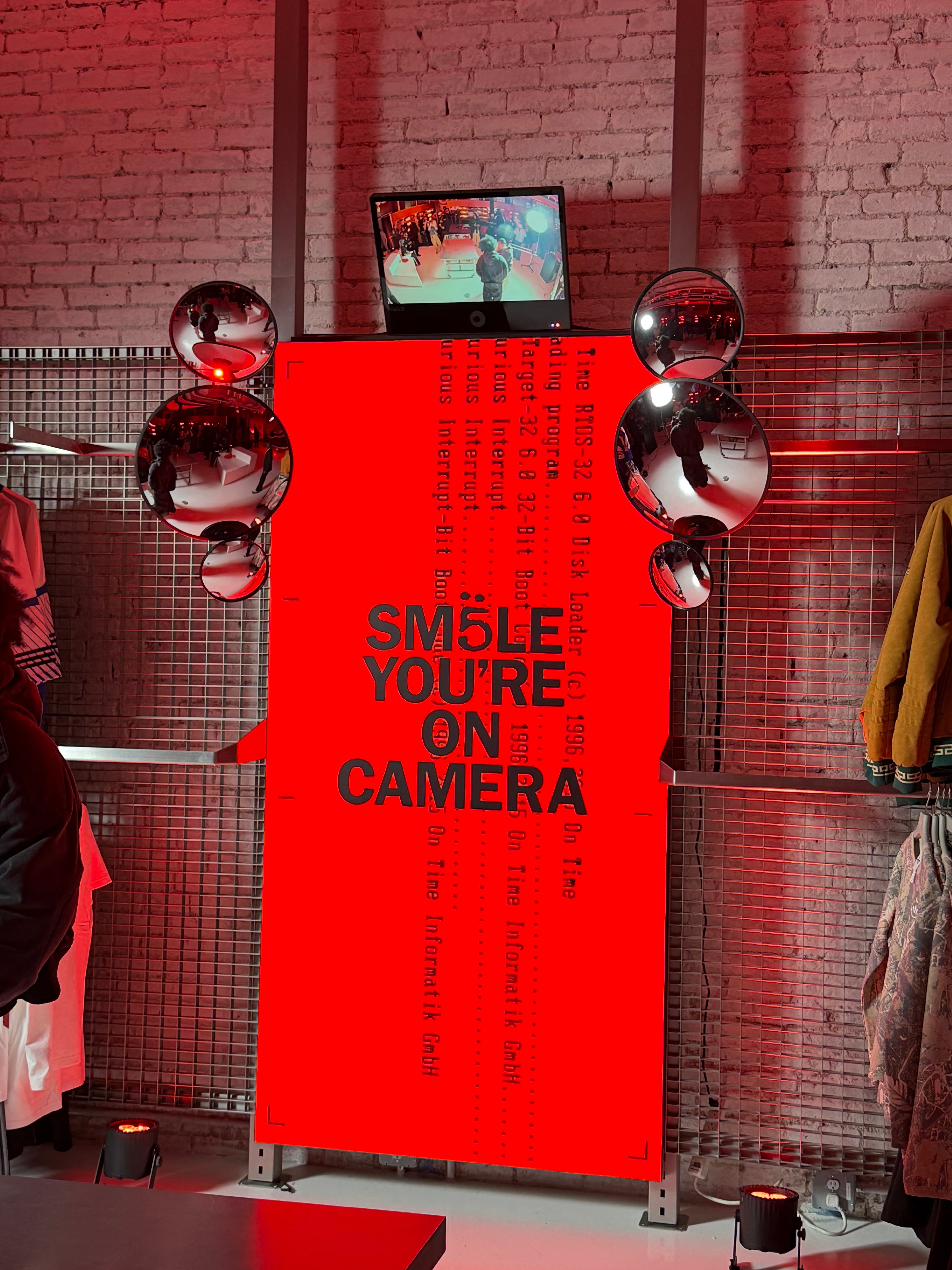 Tall red LED digital display reading 'Smile You're On Camera' with convex security mirrors in an Adidas retail store